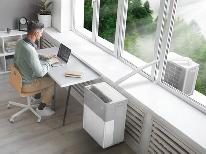 Person working at a desk with a laptop, air conditioning on the floor, and large windows showing greenery.