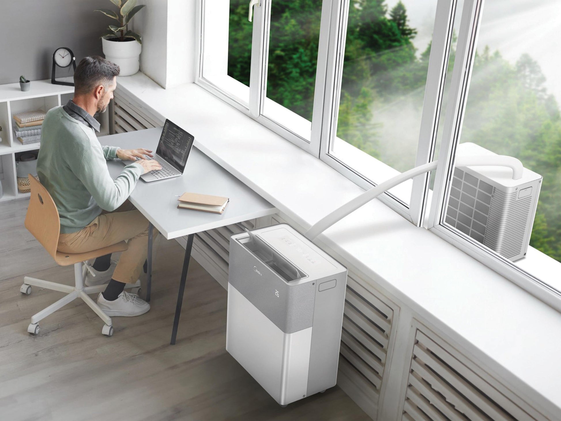 Person working at a desk with a laptop, air conditioning on the floor, and large windows showing greenery.